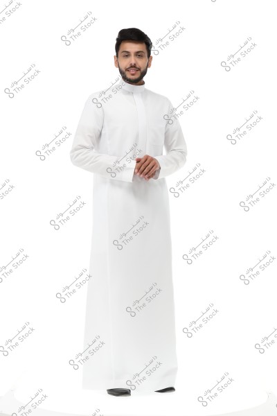 A photo of a man standing wearing a traditional white thobe, typically associated with Gulf attire, against a white background. The man is wearing black shoes and appears smiling with his hands clasped in front of him.