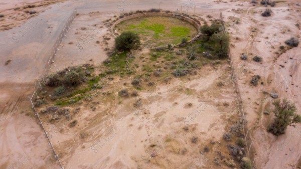 Aerial view of a desert area with a large enclosed space, featuring a circular structure partially covered with green plants. Sparse trees and shrubs are scattered across the predominantly brown desert landscape.