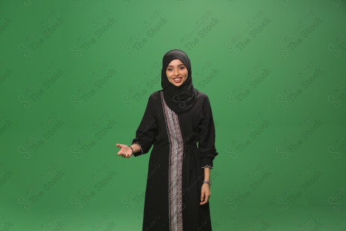 Saudi woman on green background wearing abaya and hijab, making gestures with her hands, extending her hand while smiling, Eid celebrations