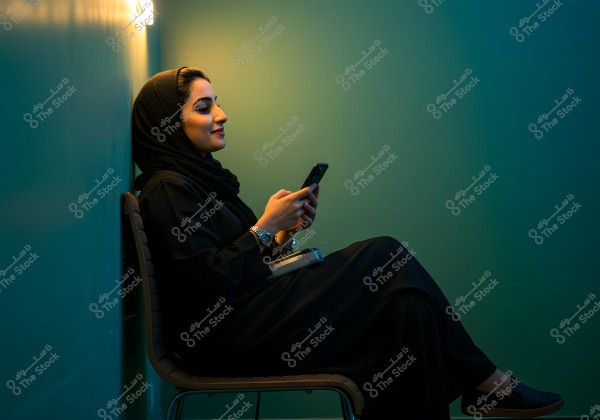 An image of a woman sitting on a chair against a blue-colored wall, wearing a black abaya and hijab, holding a mobile phone in her hands. She appears relaxed and smiling, with warm light coming from above, creating a cozy atmosphere. She wears a silver watch on her wrist and has a small notebook resting on her lap.