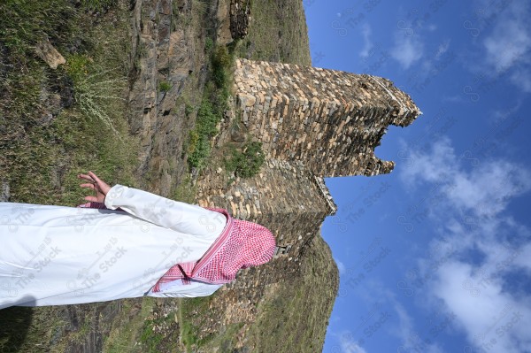 A shot of a Saudi man wearing the Saudi dress, meditating and standing on one of the green mountains.