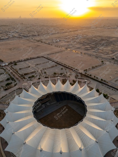 Aerial view of King Fahd Stadium in Riyadh, Saudi Arabia, at sunset. The stadium is characterized by its unique circular design and distinctive white roofs. In the background, the cityscape of Riyadh extends into the desert with the sunset casting a warm golden hue across the sky.
