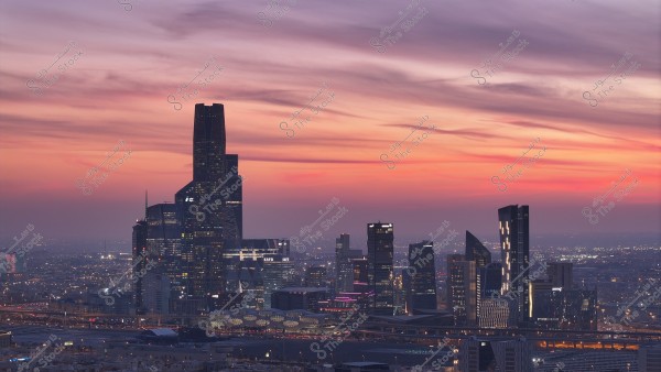 Cityscape view of Riyadh\'s skyline at sunset, with the sky painted in shades of red, orange, and purple. Several skyscrapers are visible, including a prominent tower and multiple modern buildings. The twinkling lights from the buildings add vibrancy to the city.