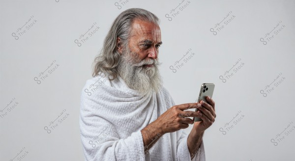 An elderly man with a long gray beard and hair, wearing a white robe, using a smartphone. He is intently focused on the device, with a plain white background.