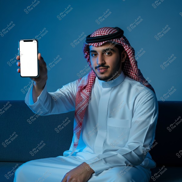 A man wearing traditional Saudi attire and a red-checkered headdress, sitting on a blue sofa. He is holding a smartphone with a blank white screen in his hand, against a light blue background.
