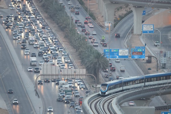 The image shows an aerial view of heavy traffic in Riyadh, Saudi Arabia. A long line of stationary vehicles is visible on the highway beside a row of palm trees. On the right side, a modern train travels along an elevated bridge. Road signs in both Arabic and English direct traffic towards destinations like King Abdulaziz Square and the Historical District. The weather appears clear with streetlights illuminating the roads.