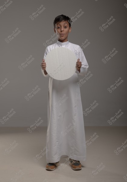 A young boy wearing a traditional white thobe stands in a studio with a light gray background. He is holding a circular white object and appears to be expressing surprise or blowing a kiss with his mouth. The boy is wearing athletic shoes.