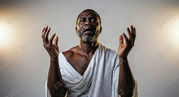 A portrait of a man standing with his hands raised in a praying position. The man is wearing a garment resembling a white Ihram. He has an expressive emotional expression, with lighting coming from both sides, giving the image a dramatic atmosphere. The background is gray.