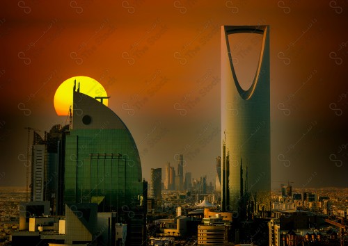 A shot showing the scheduled tower, one of the buildings and landmarks of the city of Riyadh, in front of it, a group of residential houses, and the sky is clear in orange at sunset.