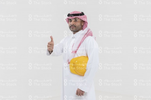 بورتريه مهندس سعودي يحمل خوذة المهندسين على يديه ويشير ويرتدي الزي السعودي الرسمي على خلفية بيضاء