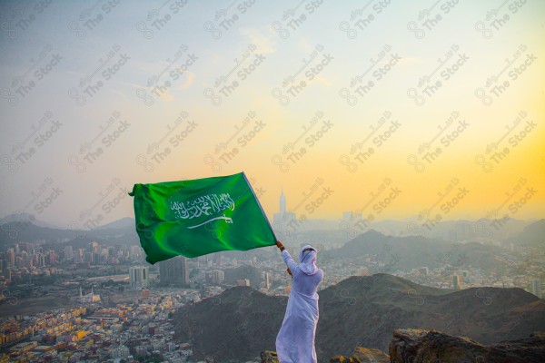 A snapshot of a Saudi man in traditional Saudi dress holding the Saudi flag over a mountain in the Makkah Al-Mukarramah region, the sky, the royal clock tower building in the Grand Mosque, buildings and landmarks, the Grand Mosque of Mecca.