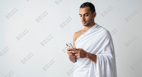A man standing against a white background, wearing a white cloth wrapped around his body, holding a smartphone and appearing engaged while looking at it.