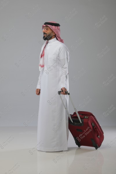 Image of a man standing in a studio against a gray background, wearing a traditional white thobe and a ghutra with a red and white checkered pattern. He is holding a red suitcase with his right hand, appearing ready for travel.