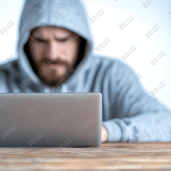 Image of a man wearing a gray hoodie sitting in front of a laptop. The man is blurred and focused on the screen, with the surface of the table in the foreground showing the main focus on the laptop.