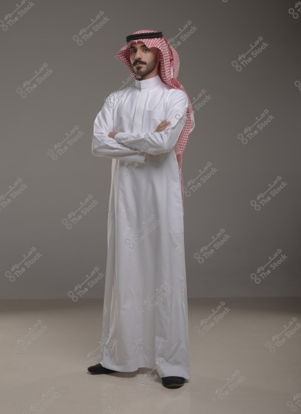Image of a man wearing a traditional Saudi thobe in white with a red shemagh, standing with his body facing forward and arms crossed. The background is a neutral gray.