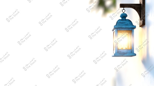 A decorative blue metallic lantern hanging from a wooden bracket on a white wall. The light glows brightly from within the lantern, casting soft shadows on the wall. Part of a tree is visible in the top left corner of the image.