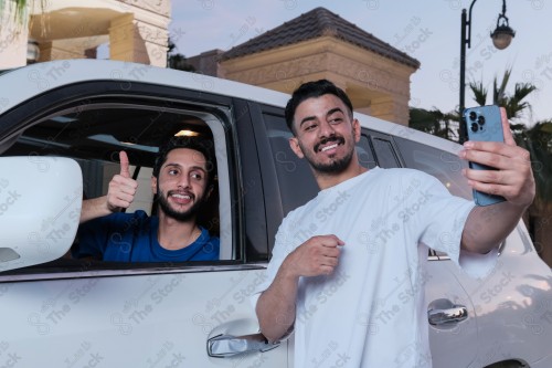 A snapshot of two young Saudis chatting and having fun on the road, their features showing joy and happiness, taking a selfie with a mobile phone