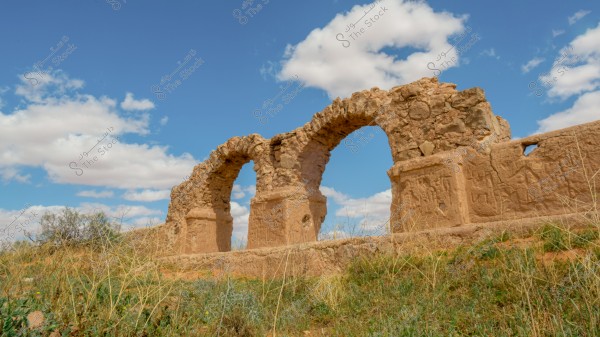 The image shows the remains of an ancient stone arch made from large, unrefined stones, surrounded by a natural setting filled with wild grasses under a blue sky dotted with scattered clouds. The image reflects ancient architectural style in a desert or semi-desert environment.