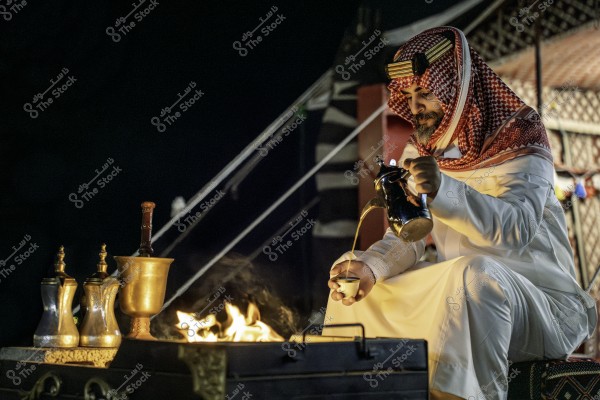 Image of a man wearing a white robe and a traditional red and white shemagh, sitting in front of a campfire. He is holding a traditional Arabic coffee pot, pouring coffee into a cup. Beside the fire, there are traditional pots and other brass containers. The image highlights a cultural scene from Arab heritage.