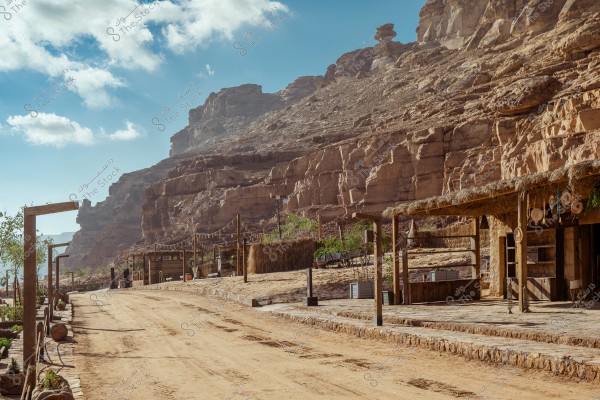 An image of a desert area with a dirt road running along a rocky cliff. Simple wooden huts with thatched roofs are on the side of the road. The environment is surrounded by natural rock formations with a blue sky and scattered clouds. There is simple decoration with hanging lights between wooden posts.
