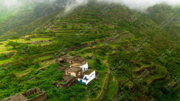 An aerial view of a green mountainous area, showcasing agricultural terraces on the mountain slopes covered with dense vegetation. A few traditional stone buildings, some with white and blue roofs, are visible amidst the lush greenery. Mist covers the mountain tops in the background, adding a sense of mystery to the landscape.