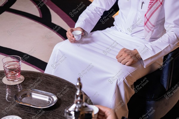 الصورة تظهر شخصًا يجلس على كرسي مرتديًا ثوبًا أبيض وغترة ب pattern حمراء وبيضاء على كتفه. الشخص يمسك بفنجان قهوة عربية ويظهر أمامه طاولة خشبية عليها كأس ماء وصينية ووعاء القهوة. السجاد ذو تصميمات هندسية بألوان مختلفة.