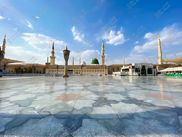 The courtyard of the Prophet\'s Mosque in Medina under a clear blue sky with scattered clouds, and the minarets of the mosque visible in the background.