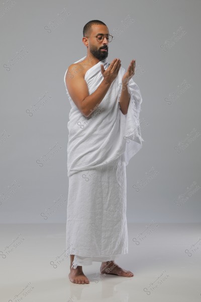 Image of a man wearing Hajj Ihram, standing in a prayer position. He is wearing glasses and has a short beard. The background is a neutral light gray.