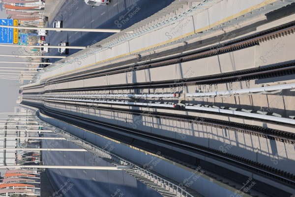 A top-down view of a train track and railway in an urban area. The track is flat and free of trains, with an adjacent highway showing some passing cars. Road signs and direction indicators are visible in the image, pointing to different areas. The weather is sunny with shadows cast along the tracks.