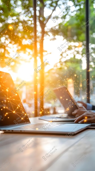A scene of an office desk featuring an open laptop on a wooden table, with a person\'s hands working on a tablet in the background. Golden glowing lights blend with the natural background of trees and leaves outside the window, creating a dynamic atmosphere.