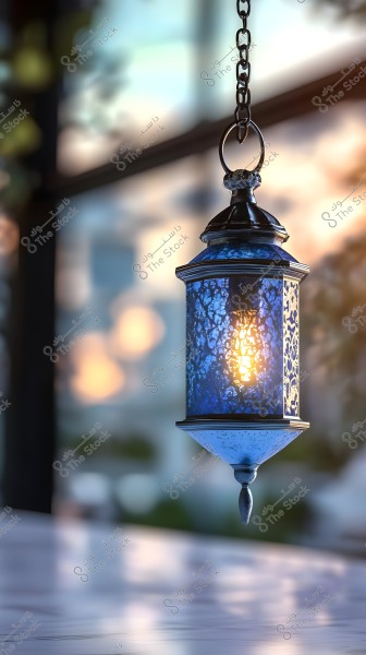 A decorative blue metal lantern hangs from a chain, glowing with warm light. The background is blurred, featuring daylight coming through a large window, creating an aesthetic effect and a tranquil ambiance.