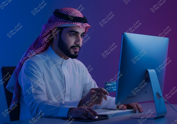 Image of a man sitting in front of a computer, wearing traditional Saudi attire with a red and white checkered headdress. He holds a bank card while typing on the keyboard. The blue and purple lighting creates a modern ambiance.