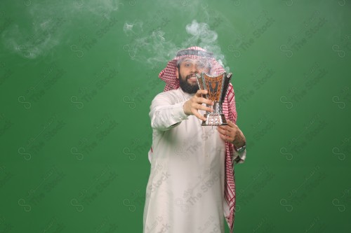 رجل سعودي يقف على خلفية خضراء يرتدي الثوب و الشماغ، يقوم بعمل حركات بيديه ، يبخر نفسه بالطيب و العود، احتفالات العيد