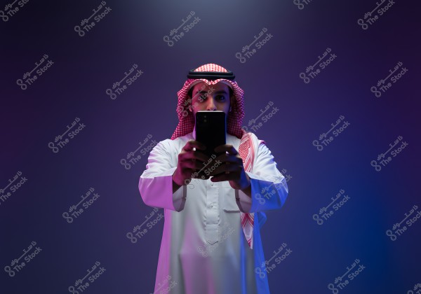 A portrait of a man wearing traditional Saudi attire with a red-checkered ghutra and a black agal. He is standing in front of a purple background, holding a smartphone centrally in the image.
