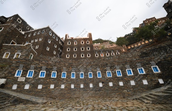 Image of a set of traditional stone-faced buildings featuring white and blue window designs. The buildings are stacked on a mountainous slope, with some trees visible in the background. The architectural design reflects the traditional culture of the Asir region in Saudi Arabia.