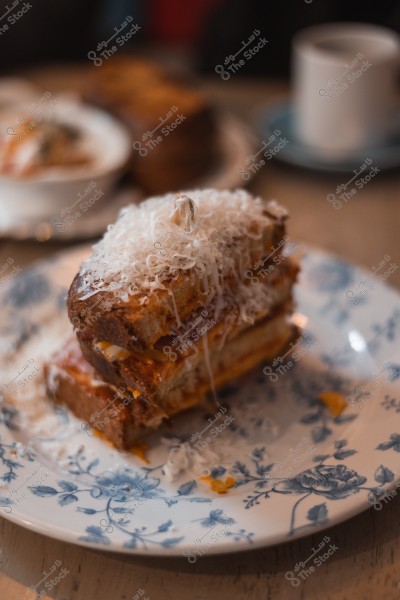 Grilled sandwich topped with shredded cheese on a decorative floral plate, with a coffee cup in the background.