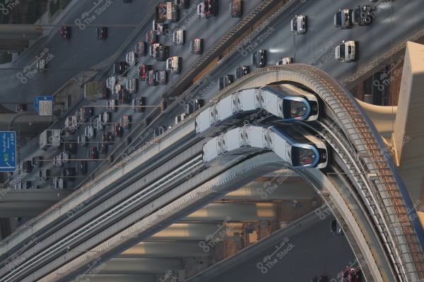 Aerial view of a metro train traveling on a bridge over a busy highway filled with cars. The road and cars are visible below as the train curves around the bridge, reflecting the sun on its metallic surface. Road signs in Arabic are visible on the side.