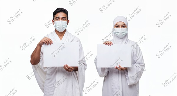 An image of a man and a woman wearing traditional Ihram clothing for Hajj and Umrah in white. Both are wearing medical face masks, standing side by side holding blank white signs. The background is white, highlighting the individuals.