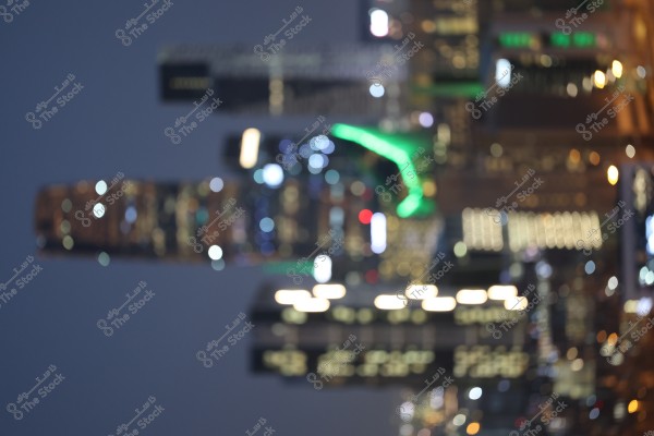 A blurry night photo of a modern cityscape with tall towers and colorful lights. The image features several skyscrapers illuminated in various colors, including a prominent green light. The background reveals a dark night sky.