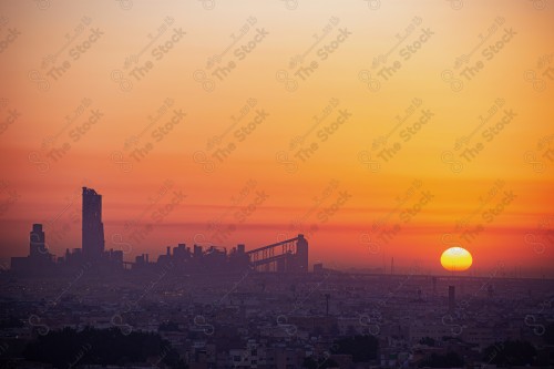 A shot showing the buildings and landmarks of the city of Riyadh, in the center of which is the cement factory. The sky appears cloudy and appears orange at sunset.