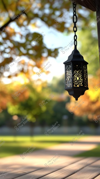 A metal lantern hanging from a chain is prominently featured on the right side of the image, with an ornate design allowing light and shadow to pass through. In the background, there is a garden with a wooden path and trees with green and yellow leaves, suggesting an autumn atmosphere.