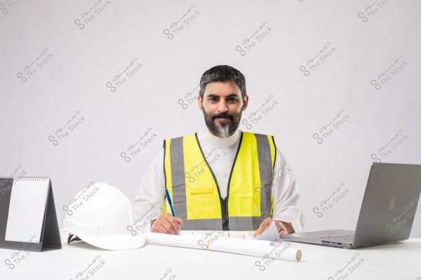 Image of a man sitting at a desk wearing a yellow reflective vest, appearing to be an engineer or construction supervisor. The man is wearing a traditional white garment, possibly from Saudi Arabia. The desk features a white helmet, a laptop, and some paper blueprints. There is also a small calendar on the desk.