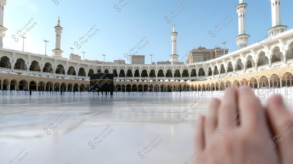 The image shows the Kaaba inside the Masjid al-Haram in Mecca, surrounded by a large number of worshippers dressed in white Ihram garments. The tall minarets and distinctive Islamic architecture of the mosque are visible in the background under a clear blue sky. In the foreground, a person\'s hands touch the ground.