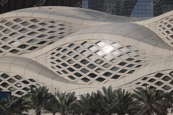 A modern architectural building with curved white panels featuring a mesh-like design, and palm trees in the foreground. The building\'s surface has distinctive curves reflecting light, and the background includes a tower with glass facades.
