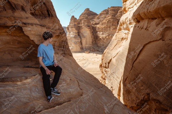 The image shows a person sitting on a rock in a mountainous desert area. The surrounding rocks are light brown and shaped by natural erosion. The person is wearing a blue t-shirt, black pants, and sneakers. The background displays tall rocky mountains under an open blue sky.