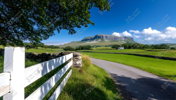 A scenic view depicting a narrow country road surrounded by expansive green fields on both sides. A white wooden fence is on the left side, and a large hill appears in the background under a clear blue sky with a few white clouds. Trees and vegetation are scattered throughout the scene.
