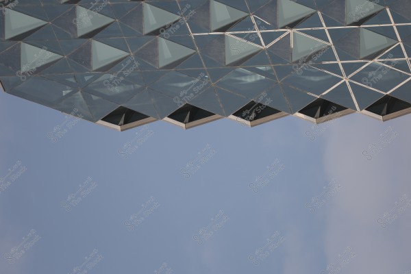 An image showing part of a modern building facade, featuring a mesh design composed of triangular glass windows pointing towards the blue sky. The structure displays its complex and symmetrical geometric details.