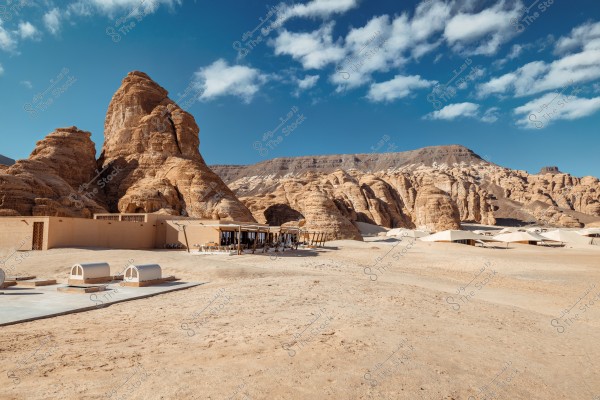 Image of a desert scene featuring natural rock formations with light brown cliffs and stones. In the foreground, there are several modern buildings covered with unique designs that blend with the surrounding nature. The sky is blue with scattered clouds.