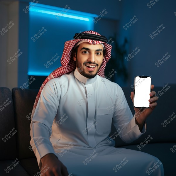 Image of a man wearing traditional white attire and a red checkered headscarf, sitting on a sofa and holding a smartphone with a blank screen. The man appears happy, with a dim blue-lit wall in the background. The image conveys a modern and relaxed atmosphere.