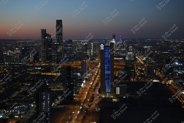 تظهر الصورة مشهداً ليلياً لمدينة ذات مبانٍ عالية مضاءة تزين الأفق. يظهر شارع طويل مضاء يتوسط المشهد ويمتد نحو الأفق. المباني تضيء بأنوار ناطحات السحاب الحديثة, مع سماء ليلية داكنة في الخلفية مرصعة بالنجوم.